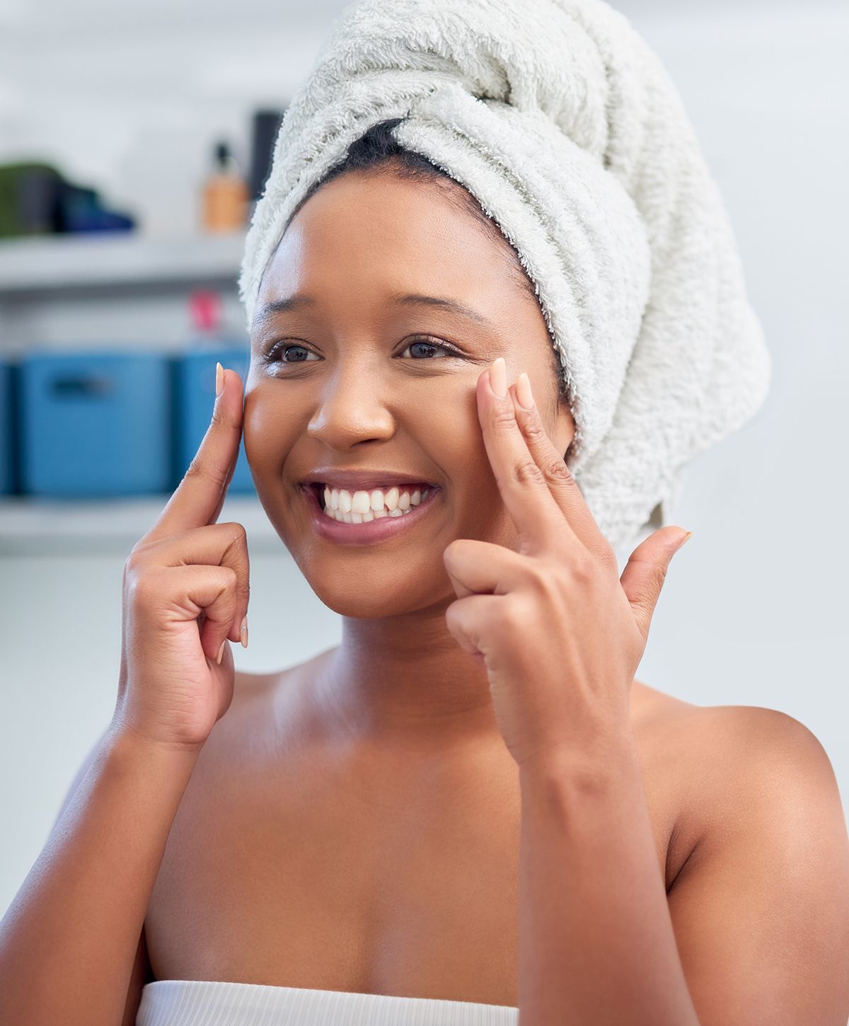 Smiling woman applying skincare with towel on head.